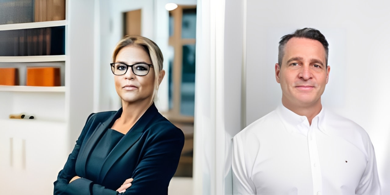 Zwei professionell gekleidete Personen stehen in einem modernen Büro, die Frau trägt Brille und hat kurze blonde Haare, der Mann hat dunkle, kurze Haare.