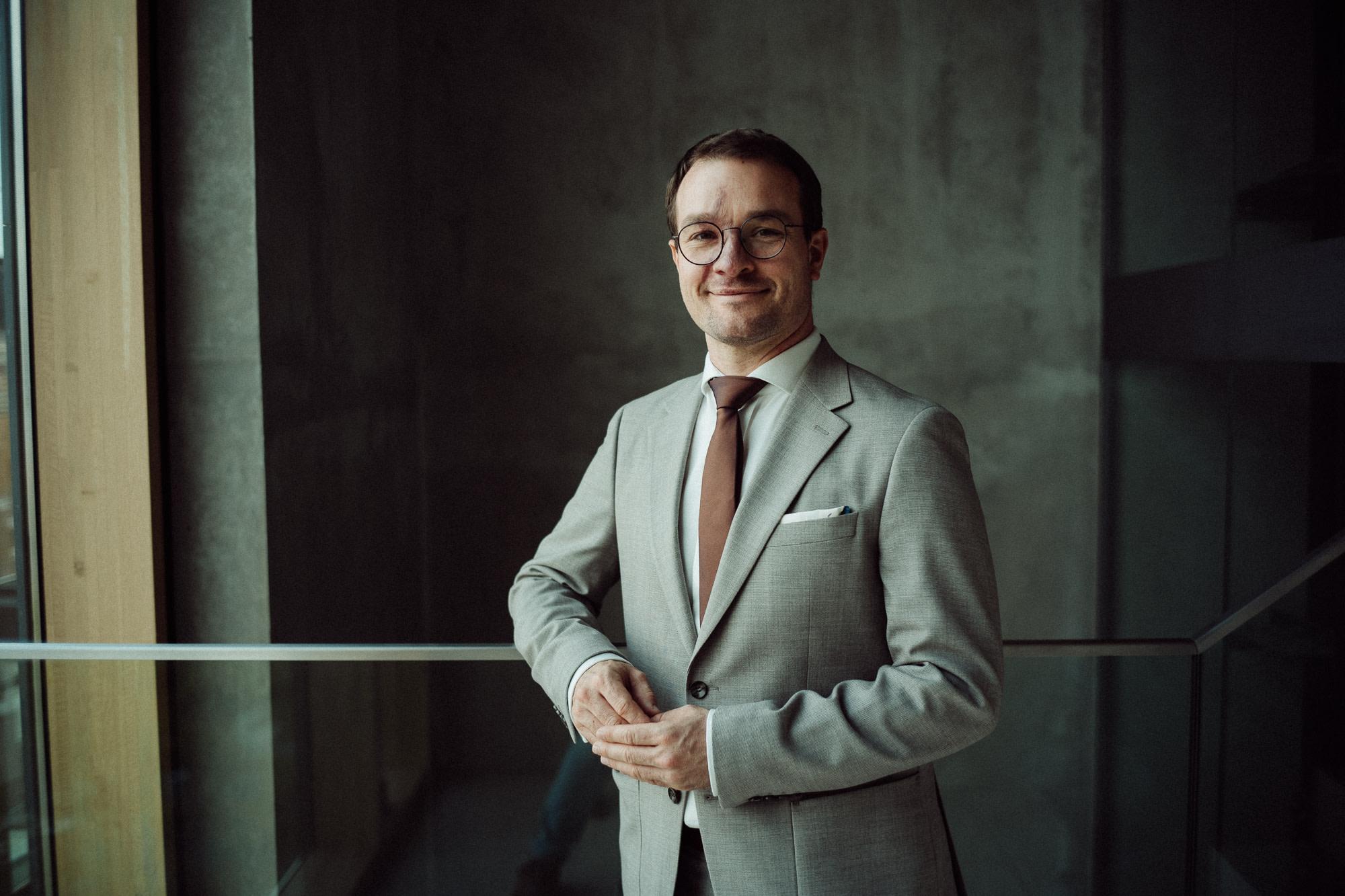 Geschäftsmann in einem hellgrauen Anzug mit Brille, der an einer Glastür steht.