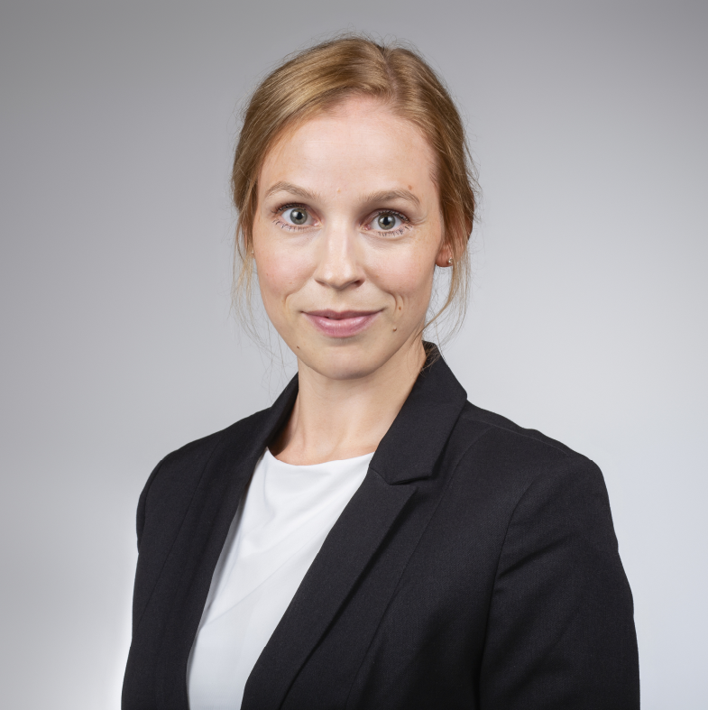 Porträt einer professionellen Frau mit blonden Haaren, die einen schwarzen Blazer und ein weißes Hemd trägt, vor einem grauen Hintergrund.