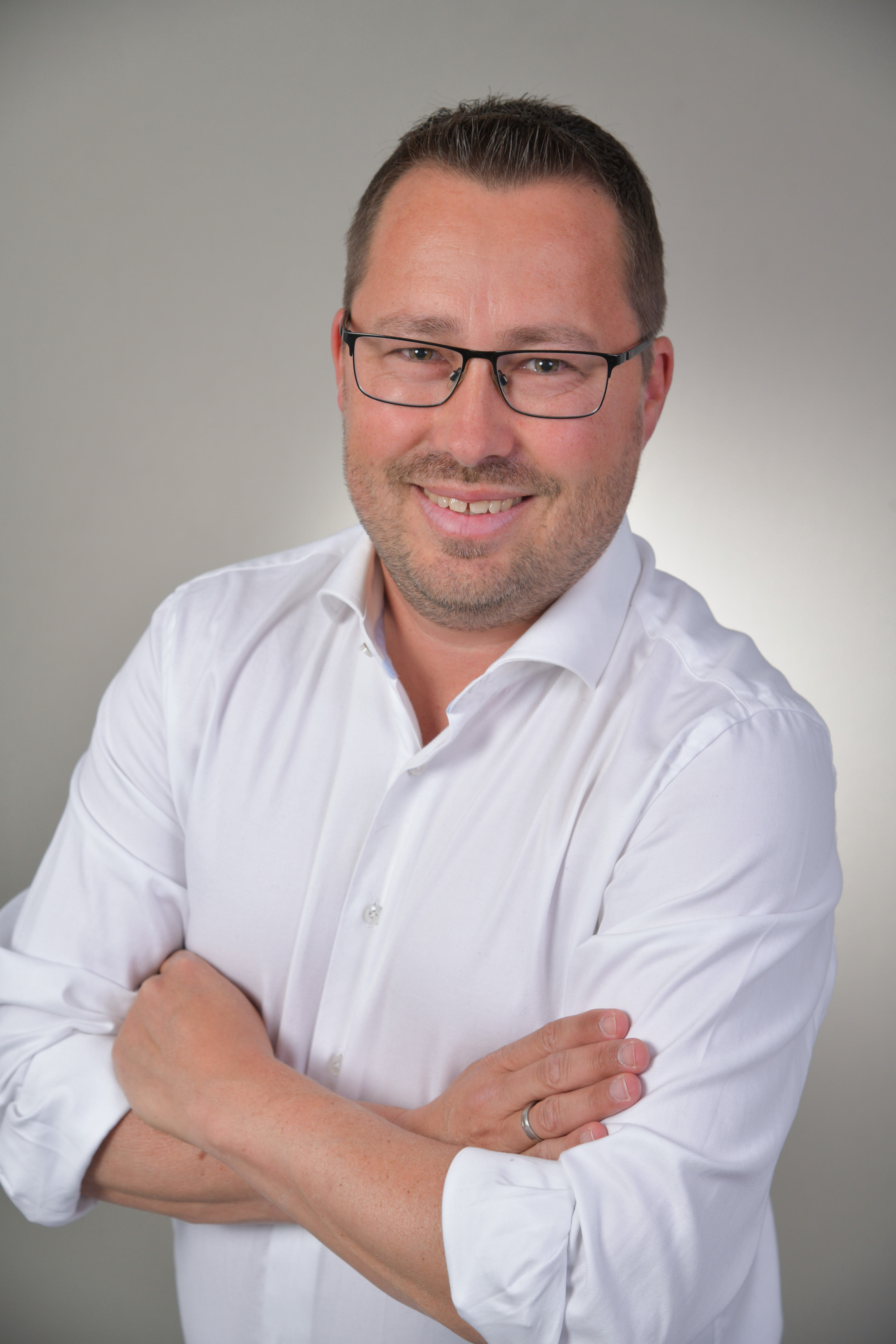 Lächelnder Mann mit Brille in weißem Hemd, Arme verschränkt, vor neutralem Hintergrund.