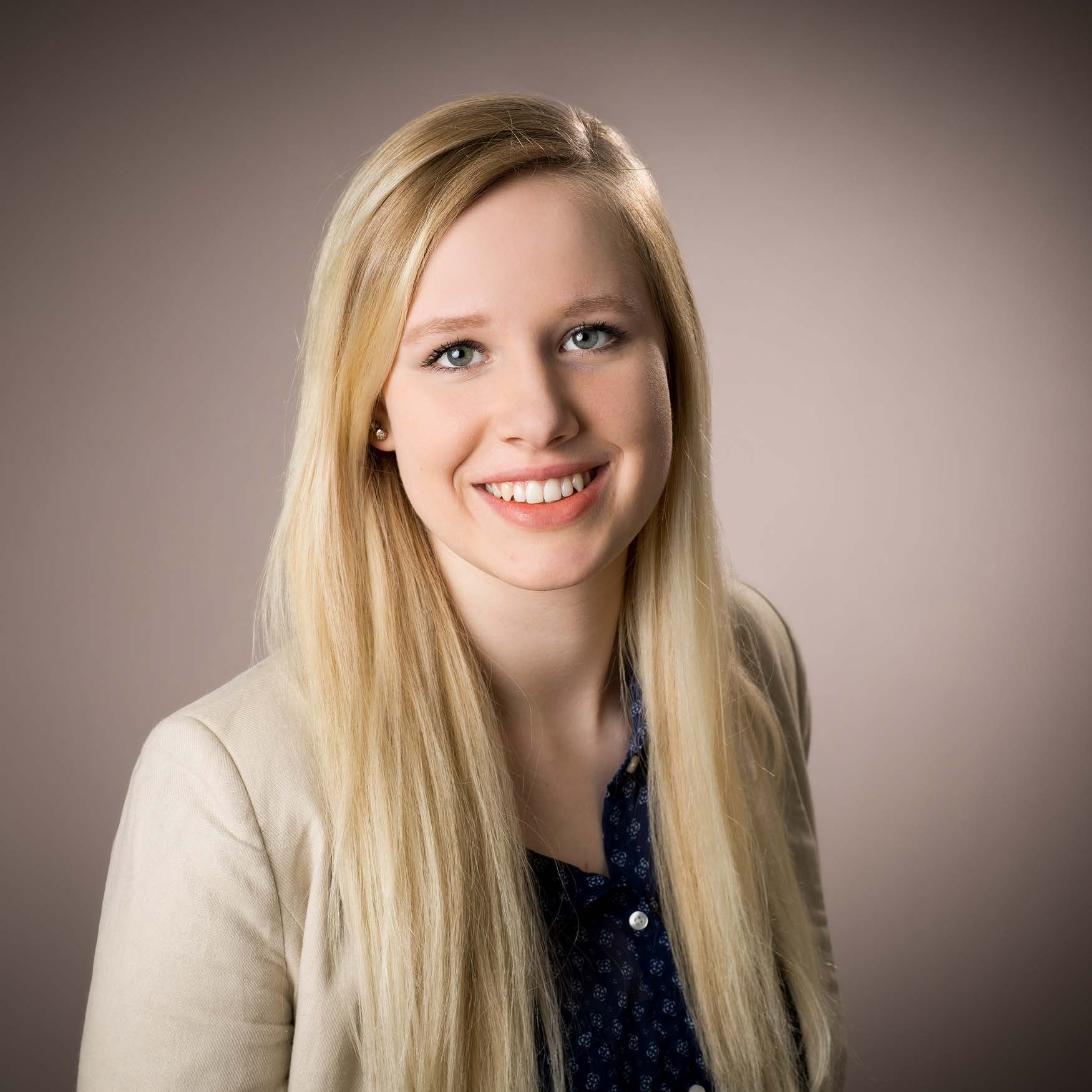 Porträt einer jungen Frau mit langen blonden Haaren, blauen Augen und einem freundlichen Lächeln, trägt eine beige Jacke und eine dunkelblaue Bluse.