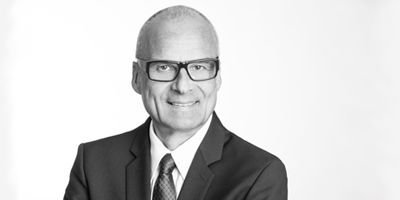 Porträt eines Mannes in formeller Kleidung mit Brille, lächelnd, in Schwarz-Weiß.