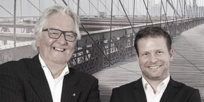 Zwei Männer in formeller Kleidung stehen auf einer Brücke mit einer Hängebrücke im Hintergrund.