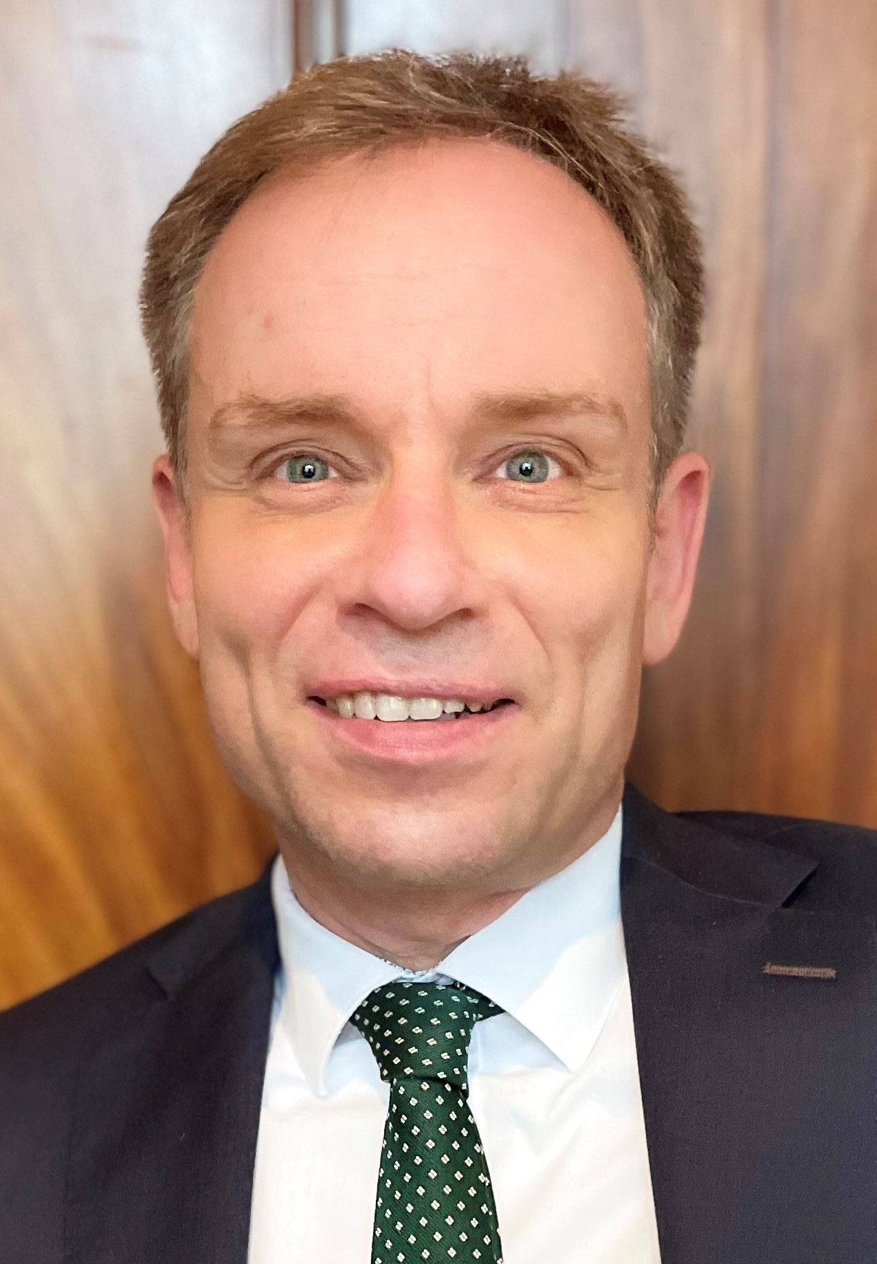 Porträt eines Mannes in formeller Kleidung vor einer Holzwand.