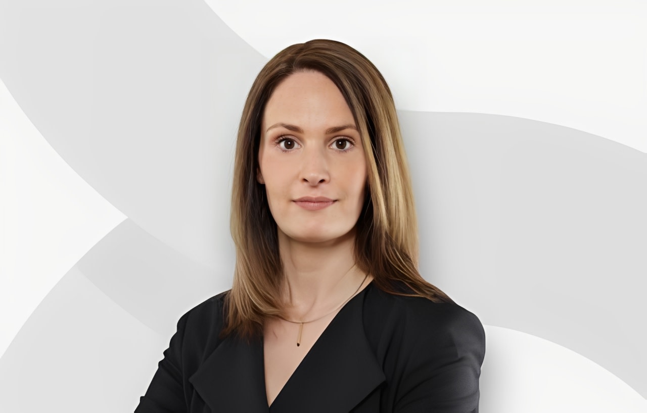 Porträt einer jungen Frau mit braunen Haaren, schwarzer Kleidung und dezentem Schmuck vor einem hellen, abstrakten Hintergrund.