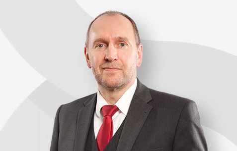 Ein Mann in einem dunklen Anzug mit roter Krawatte vor einem hellen Hintergrund.