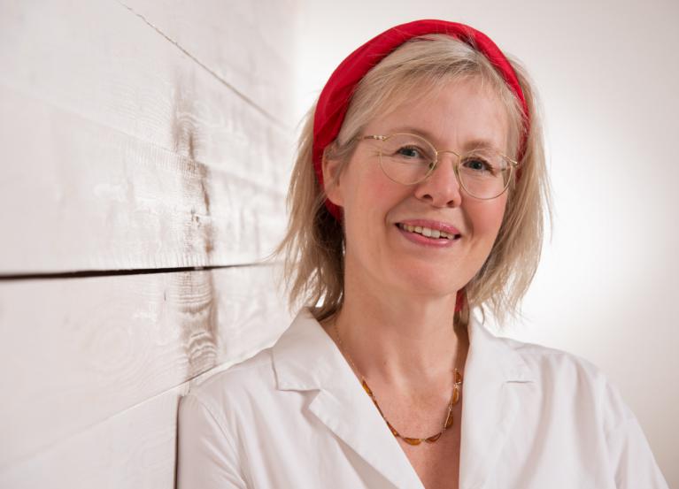 Lächelnde Frau mit Brille, rotem Haarband und weißem Kittel vor einer Holzwand.