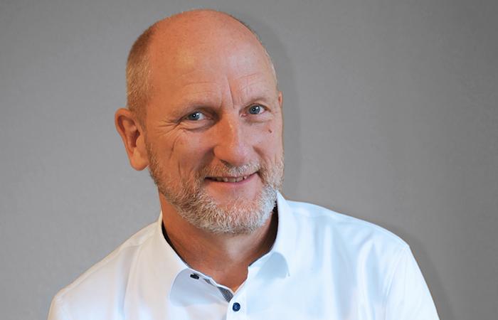 Ein lächelnder Mann mit kurzem, grauem Bart und blauen Augen, trägt ein weißes Hemd und steht vor einem grauen Hintergrund.