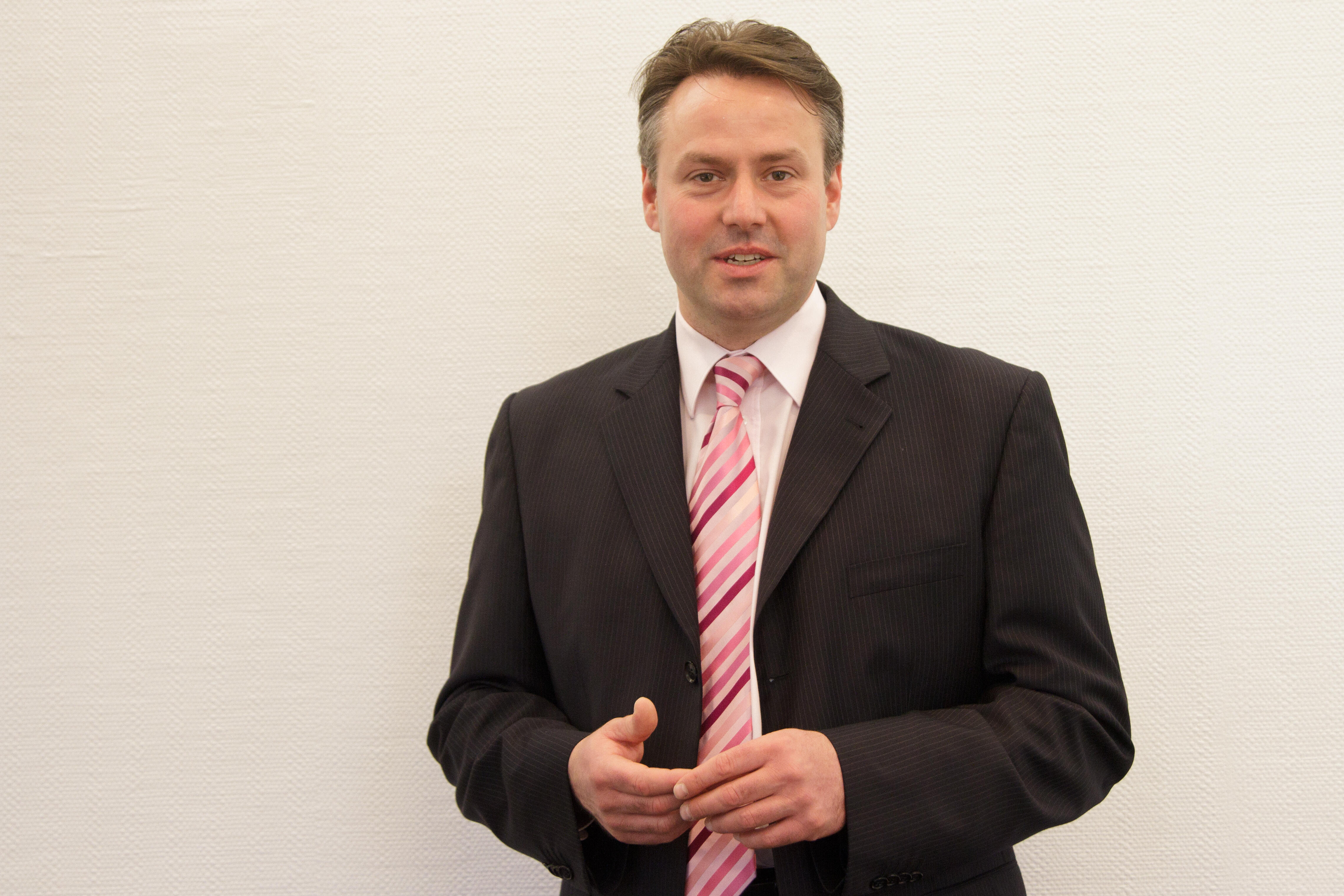 Ein Mann in einem schwarzen Anzug mit rosa gestreiftem Krawatte vor einer weißen Wand.