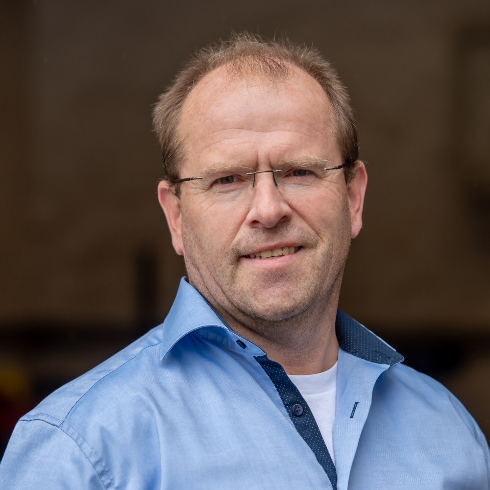 Mann mit Brille und blauer Hemd, lächelnd, im Freien.