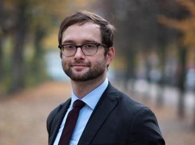 Ein Mann in formeller Kleidung mit Brille, im Freien mit herbstlichen Bäumen im Hintergrund.