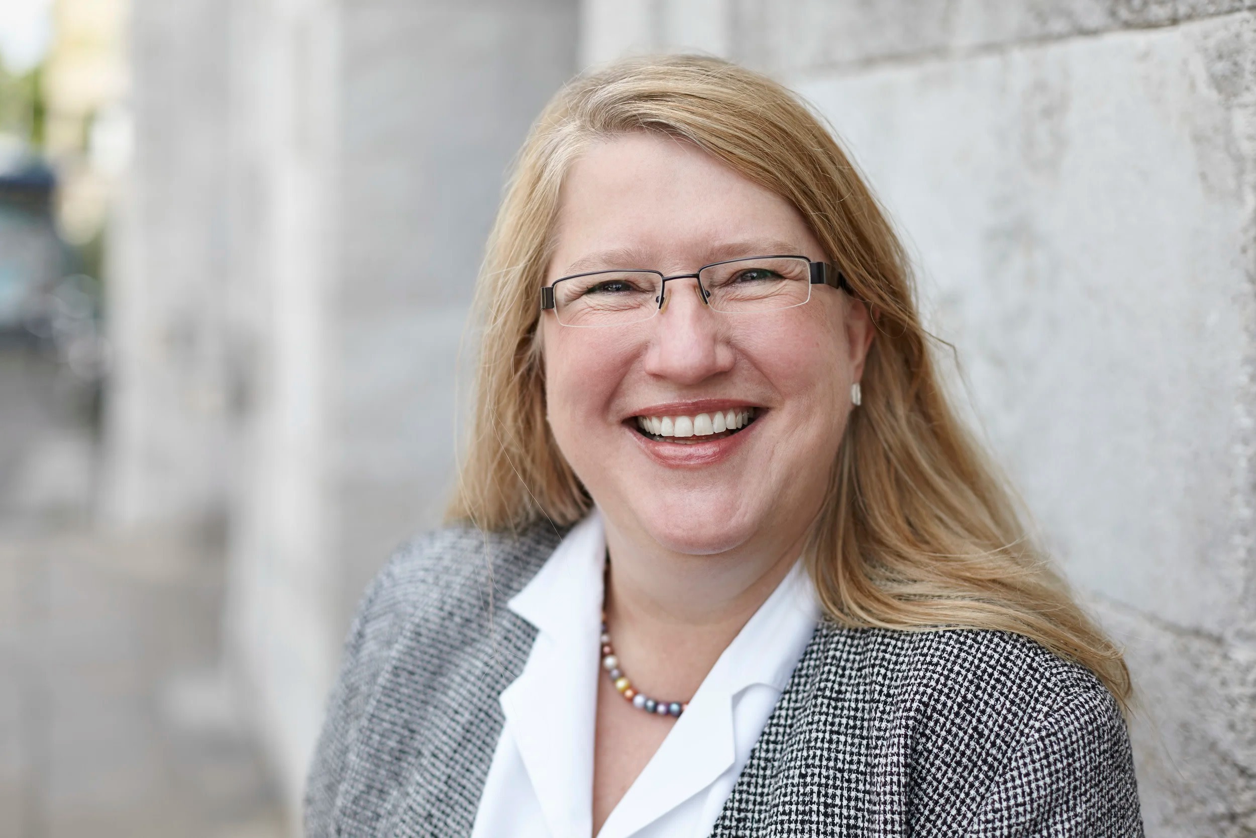 Lächelnde Frau mit Brille vor einer Steinmauer.
