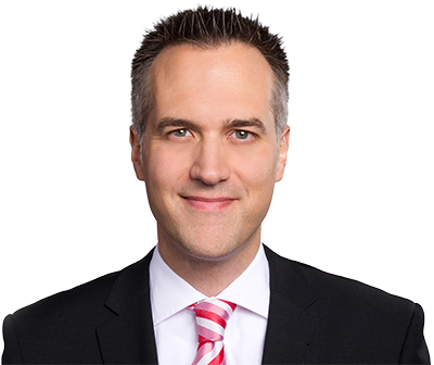 Ein professionelles Porträt eines Mannes in formeller Kleidung, der einen schwarzen Anzug, ein weißes Hemd und eine rosa-weiße Krawatte trägt.