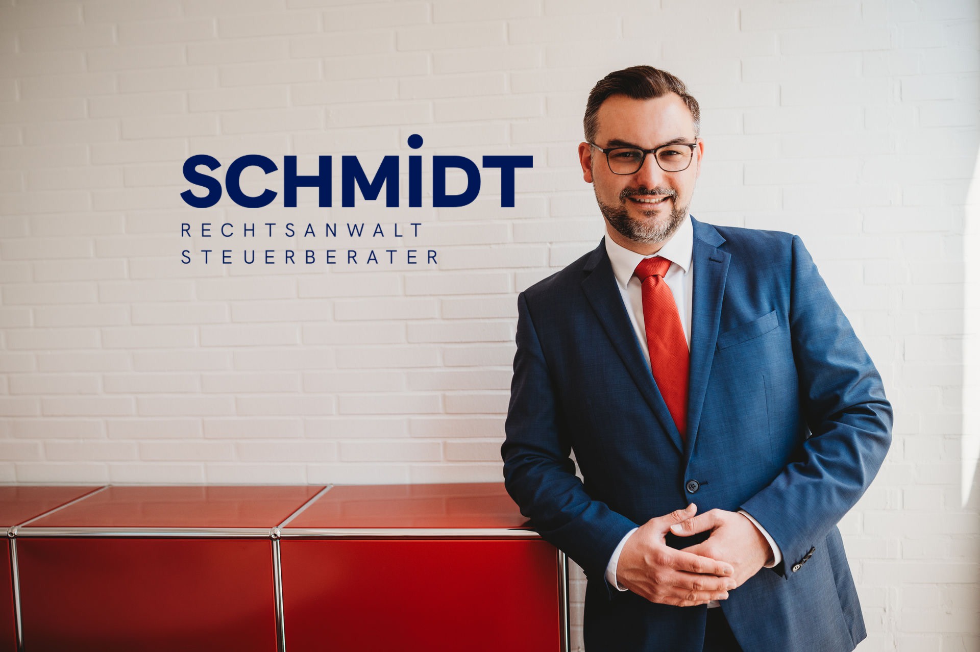 Ein Mann in Anzug mit roter Krawatte steht vor einer weißen Ziegelwand mit dem Text "SCHMIDT RECHTSANWALT STEUERBERATER" auf der linken Seite.