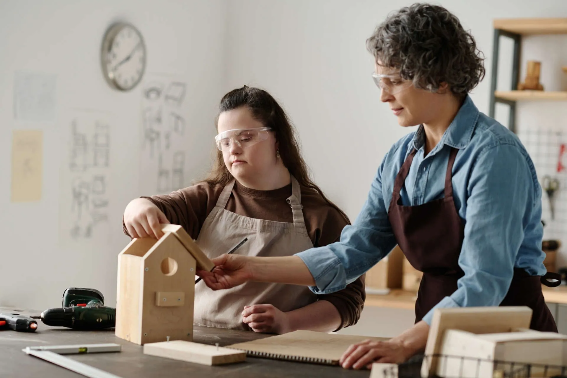 Eine junge Frau und eine ältere Frau bauen gemeinsam ein Vogelhaus in einer Werkstatt.