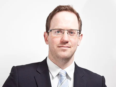 Porträt eines Mannes in formeller Kleidung mit Brille vor hellem Hintergrund.