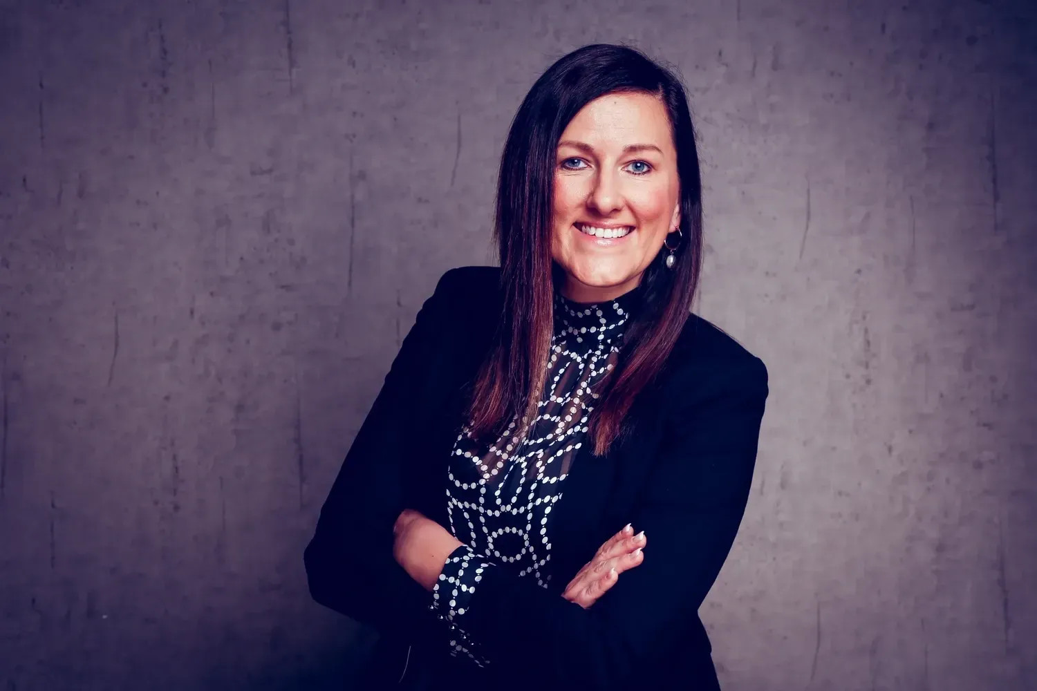 Frau in schwarzer Jacke und polka-dot Bluse, lächelnd, vor grauer Wand.