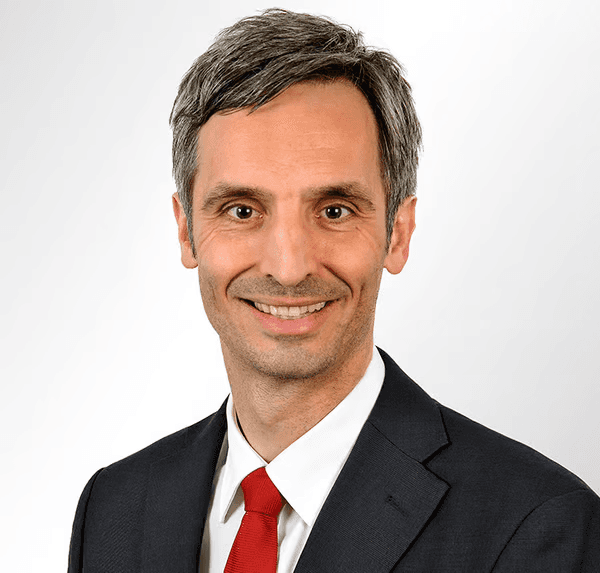 Porträt eines lächelnden Mannes in Anzug mit roter Krawatte vor weißem Hintergrund.