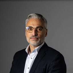 Porträt eines Mannes mit grauem Haar und Brille, in formeller Kleidung, vor grauem Hintergrund.