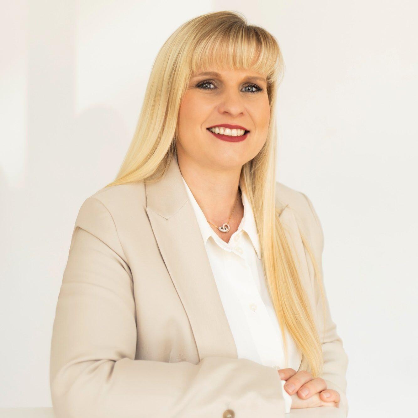 Porträt einer lächelnden Frau mit langen blonden Haaren, in einem weißen Hemd und beigem Blazer, vor hellem Hintergrund.