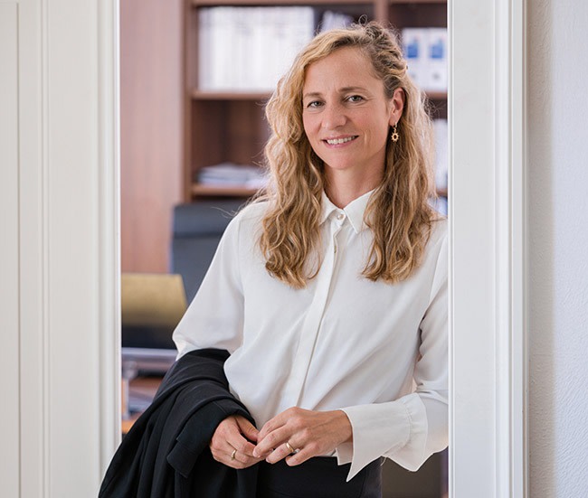 Frau steht in einem Büro, lächelnd, mit einem schwarzen Mantel in der Hand.