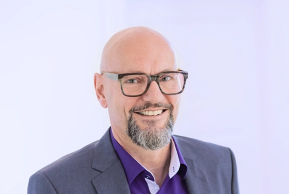 Ein lächelnder Mann mit Brille, Glatze und Bart, trägt einen dunklen Anzug und eine lila Hemd.