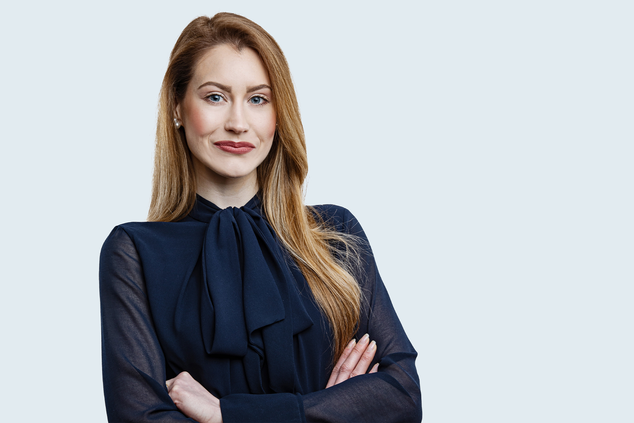 Selbstbewusste Frau mit roten Haaren in dunklem Bluse mit Schleife, die die Arme verschränkt.