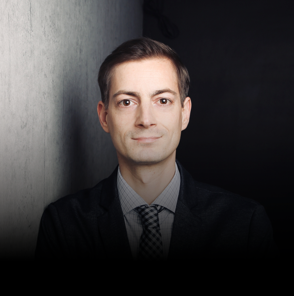 Porträt eines Mannes in formeller Kleidung vor einer dunklen Wand.