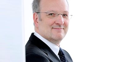 Geschäftsmann in Anzug mit Brille, lächelnd, vor hellem Hintergrund