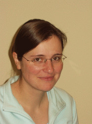 Porträt einer Frau mit dunklem, kurzem Haar und Brille, lächelnd vor einem hellen Hintergrund.
