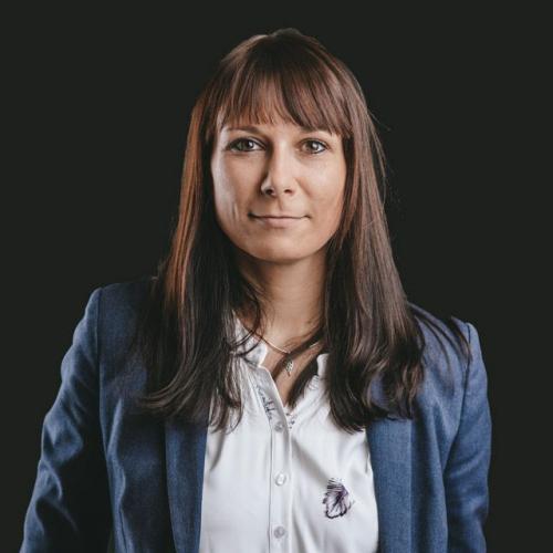Eine Frau mit braunen Haaren und geraden Frisur, trägt eine weiße Bluse und eine blaue Jacke, vor dunklem Hintergrund.