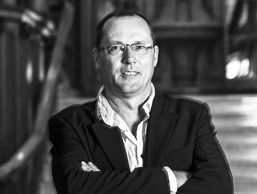 Mann in formeller Kleidung mit Brille, armselig, vor einem holzverkleideten Hintergrund.