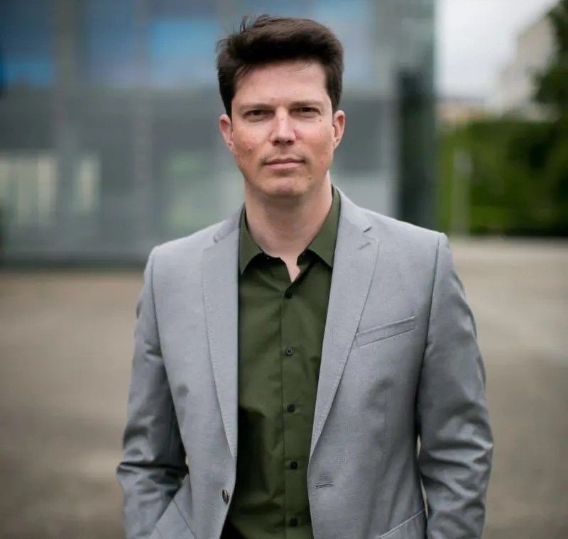 Mann in grauem Anzug und grüner Hemd vor einem modernen Gebäude.