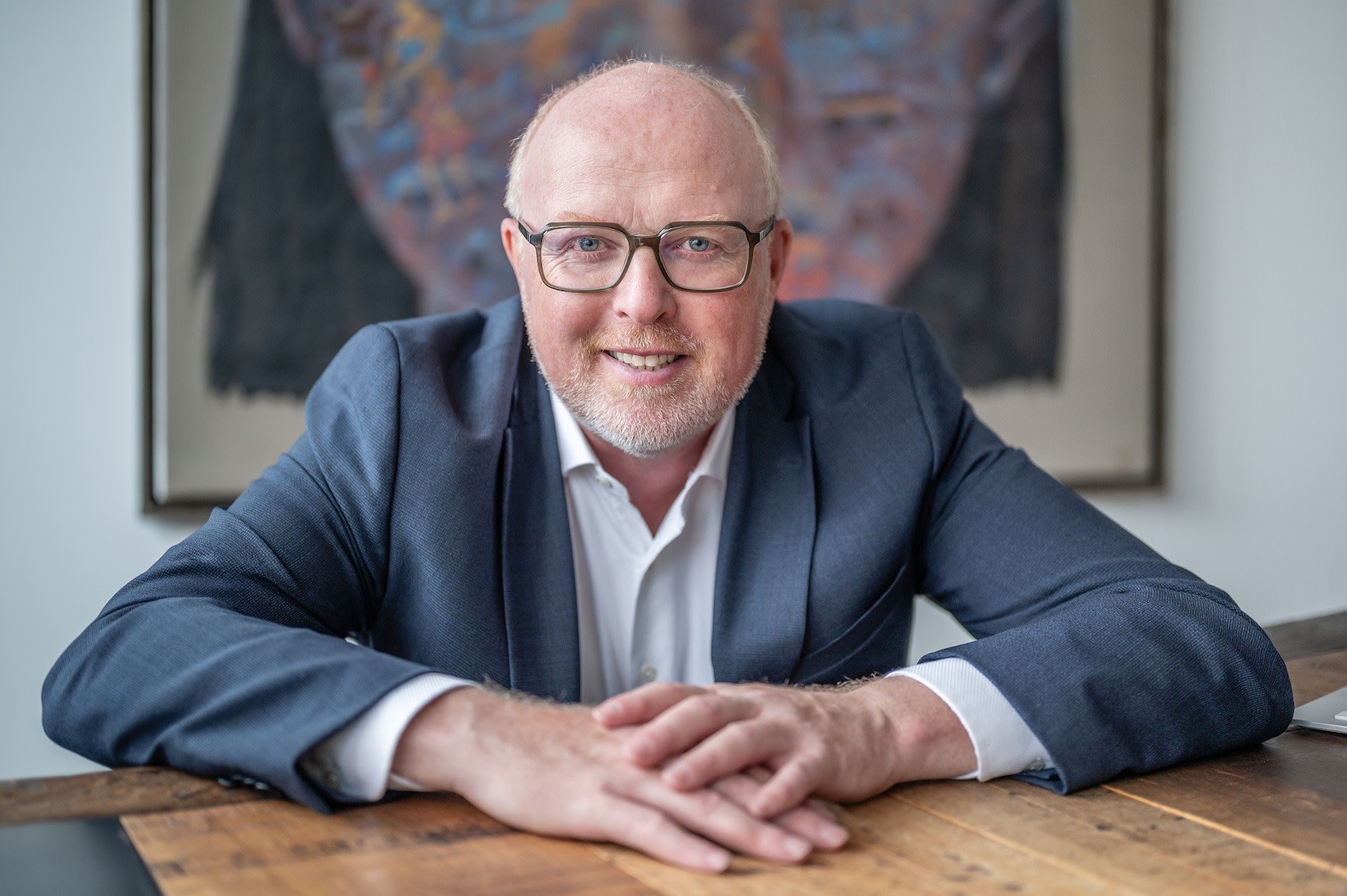 Lächelnder Mann in Brille und Anzug sitzt an einem Holztisch.