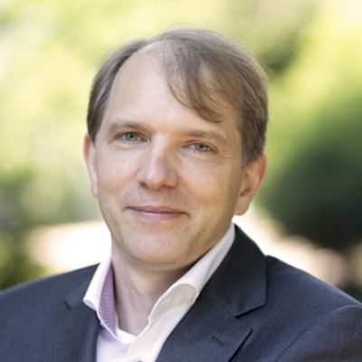 Porträt eines Mannes in einem dunklen Anzug vor einem grünen, unscharfen Hintergrund.