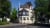 Mehrstöckiges Haus mit dunklem Dach und weißen Fenstern, umgeben von Bäumen, an einer Straßenecke in einer Stadt.