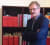 Ein Mann mit Brille steht vor einem Bücherregal mit roten und weißen Büchern, die er anlächelt.