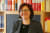 Frau mit Brille und dunklem Blazer vor einem Bücherregal mit bunten Büchern.